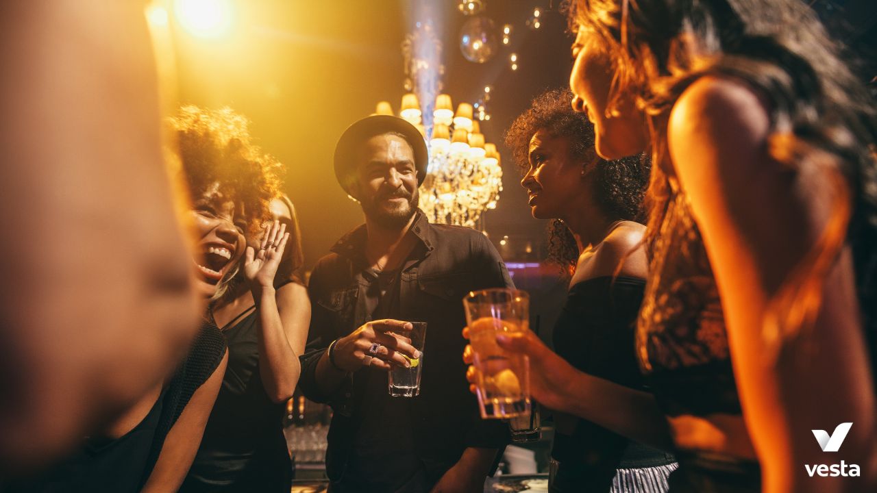 A group of young adults socializing at a dimly lit nightclub. A man in a black outfit and hat smiles while holding a drink, surrounded by women engaged in conversation and laughter. Warm lighting and chandeliers add to the vibrant atmosphere. The "Vesta" logo is visible in the bottom right corner.