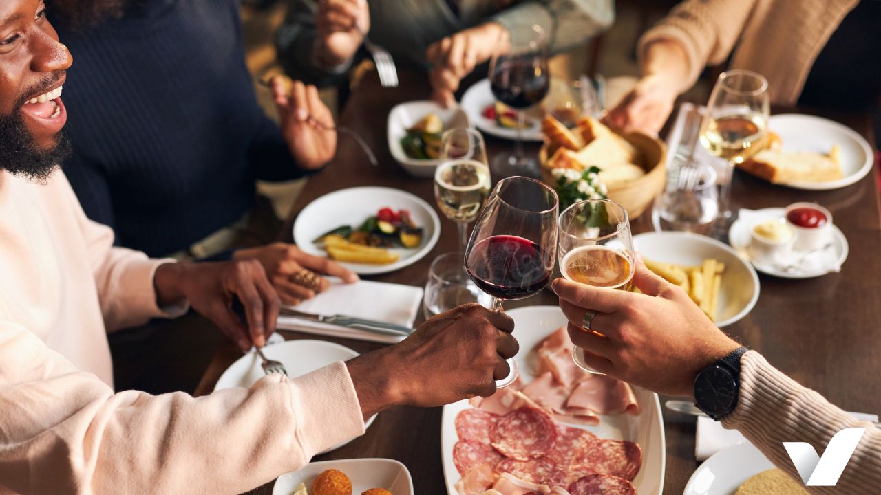 A group of friends enjoys a meal together at a restaurant event, raising glasses of red wine and beer in a toast. The table is filled with various dishes, including a charcuterie plate, fries, bread, and glasses of white wine. The atmosphere is warm and lively, with people smiling and engaging in conversation.