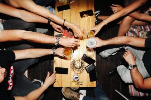 op-down view of several people clinking glasses of beer and cocktails over a wooden table at a bar or social gathering. The table is cluttered with smartphones, wallets, and a small candle centerpiece. One person is wearing a wristwatch, and a striped cushion and cat are visible on the side.
