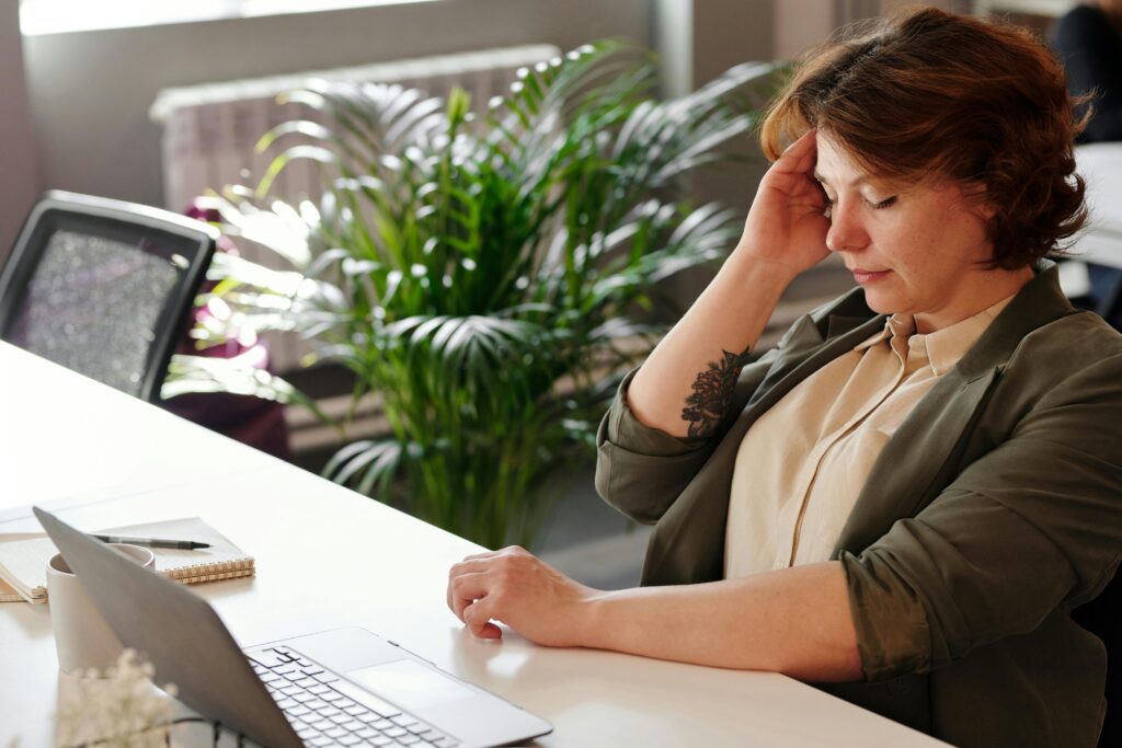 Woman sitting at a desk with her eyes closed and hand on her forehead, appearing stressed or fatigued from posting event listings to dozens of local calendars. A laptop, notebook, and coffee cup are on the desk, and a large green plant is in the background.