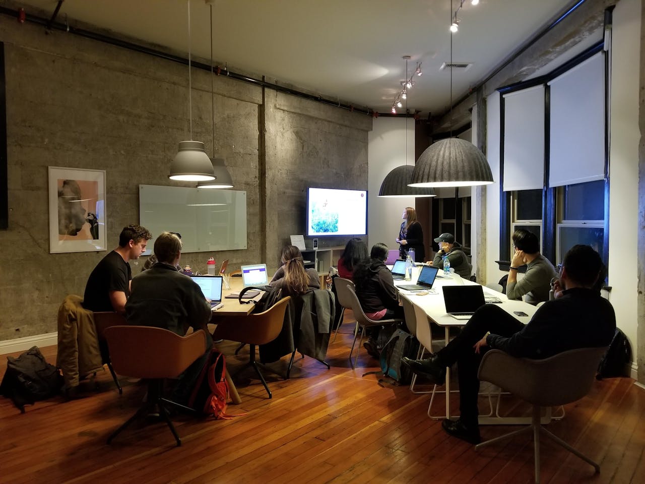 A news editorial team seated around a long conference table in a modern office, giving press release feedback. Most attendees have laptops open, and the room has exposed concrete walls, large pendant lights, and wood flooring.